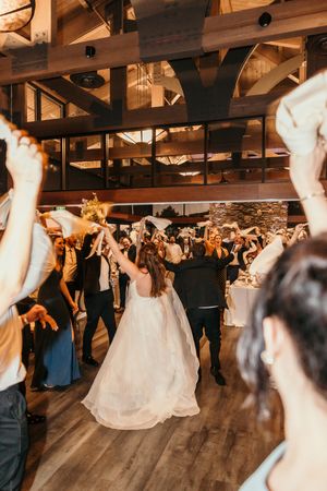 Bride and groom walk through cheering guests waving napkins at indoor wedding reception on wooden dance floor.