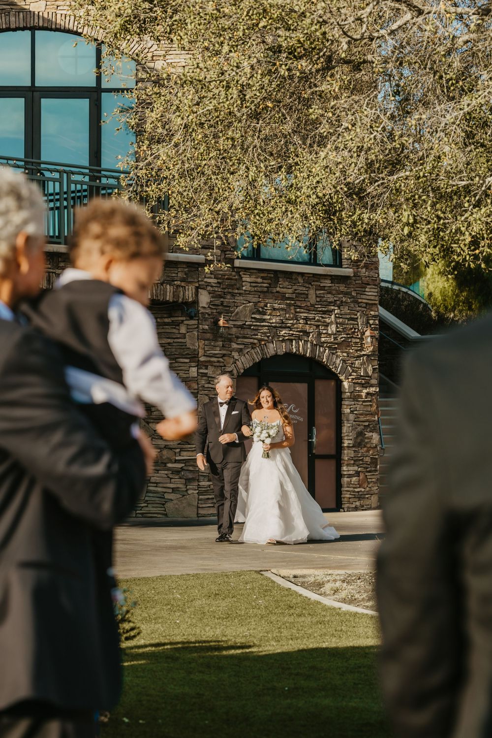 Bride walks down outdoor aisle with escort past stone building and tree-lined venue during sunny wedding ceremony.