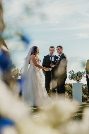 Bride and groom exchange vows outdoors with officiant, flanked by blue and white floral arrangements under a bright sky.