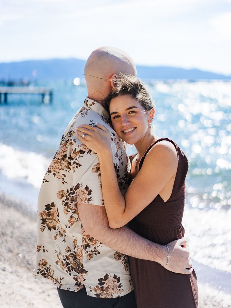 Couple embracing on a sunny beach near a lake, woman smiling in brown dress, man in floral shirt.