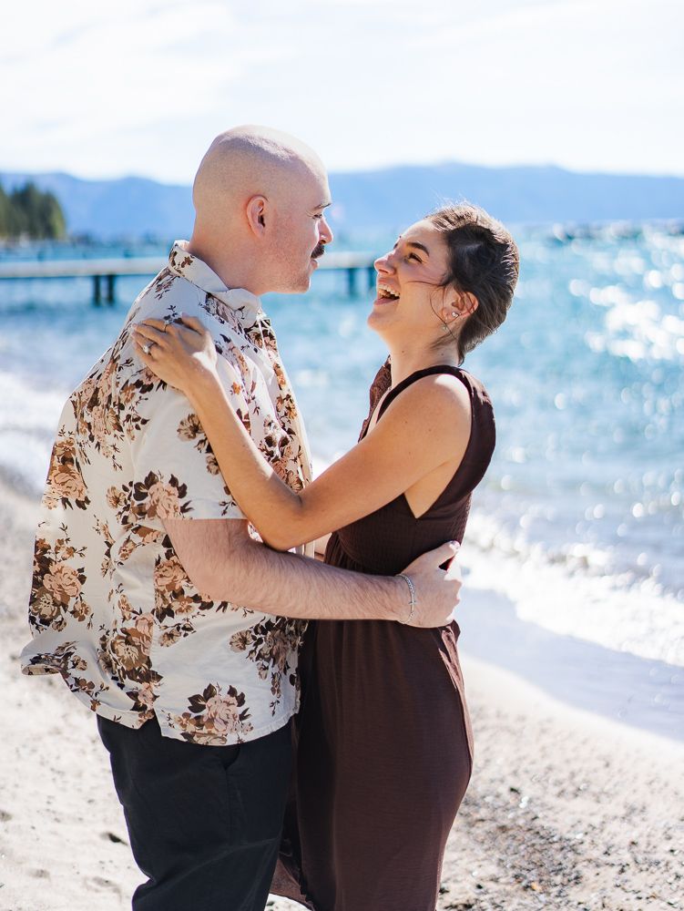 Happy couple embracing and laughing on a sunny beach near Lake Tahoe, man in floral shirt, woman in brown dress.