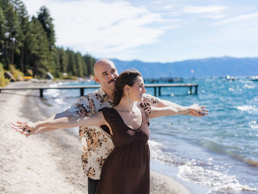 Couple posing joyfully on Lake Tahoe beach with arms outstretched, mountains and pier in background.