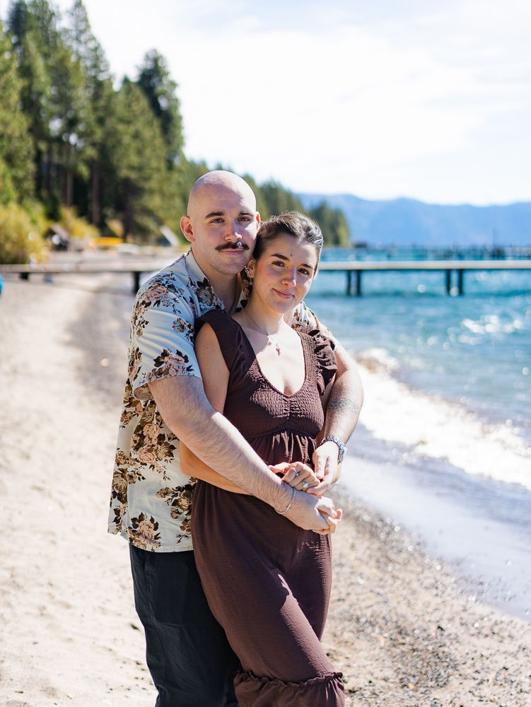 Couple embracing on a rocky beach at Lake Tahoe, with turquoise water, a pier, and pine-covered mountains in the background.