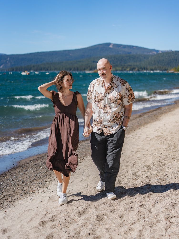 Couple walking hand-in-hand on a sunny beach with blue lake and mountain views in the background.