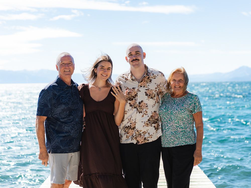 Family of four smiling together on a dock with a scenic blue ocean and mountains in the background.