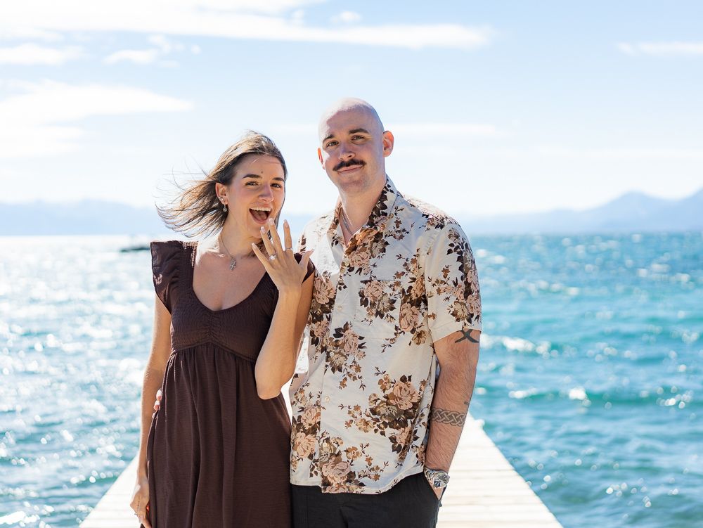 Happy couple on a dock showing off engagement ring with turquoise lake and blue sky in the background.