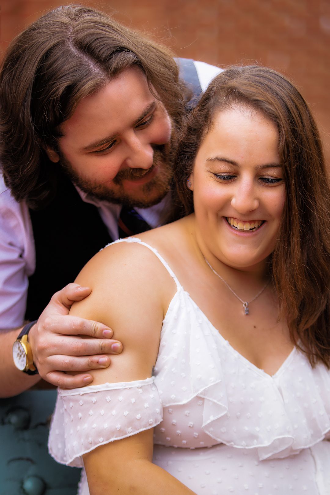 A smiling couple in wedding attire share a tender moment, man in vest embraces woman in white off-shoulder dress.