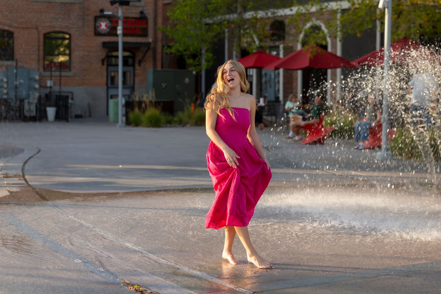 senior girl wearing a pink dress barefoot splashing around on a splash pad