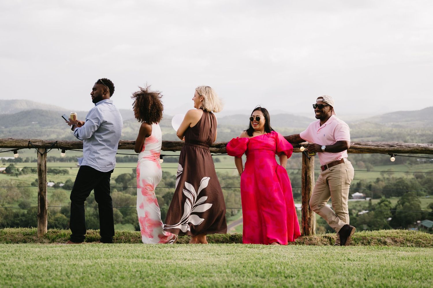 Five friends in colorful formal attire gather at a wooden fence overlook with scenic green hills and mountains behind them.
