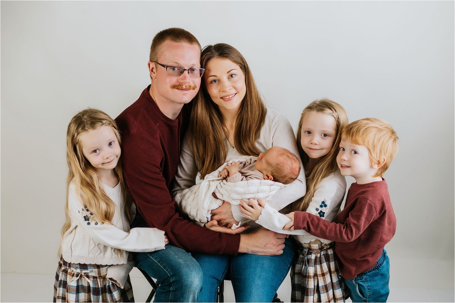 A happy family of six poses together, parents holding a newborn baby surrounded by three young children in a bright studio.