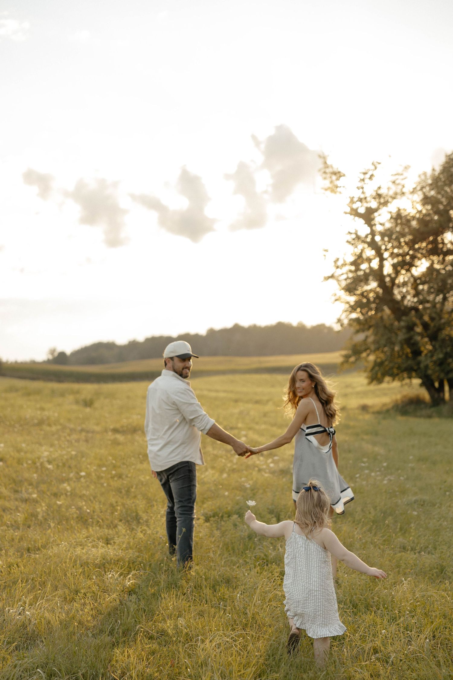 Family of three holding hands in a sunny green field at golden hour, with a young girl in a white dress twirling.