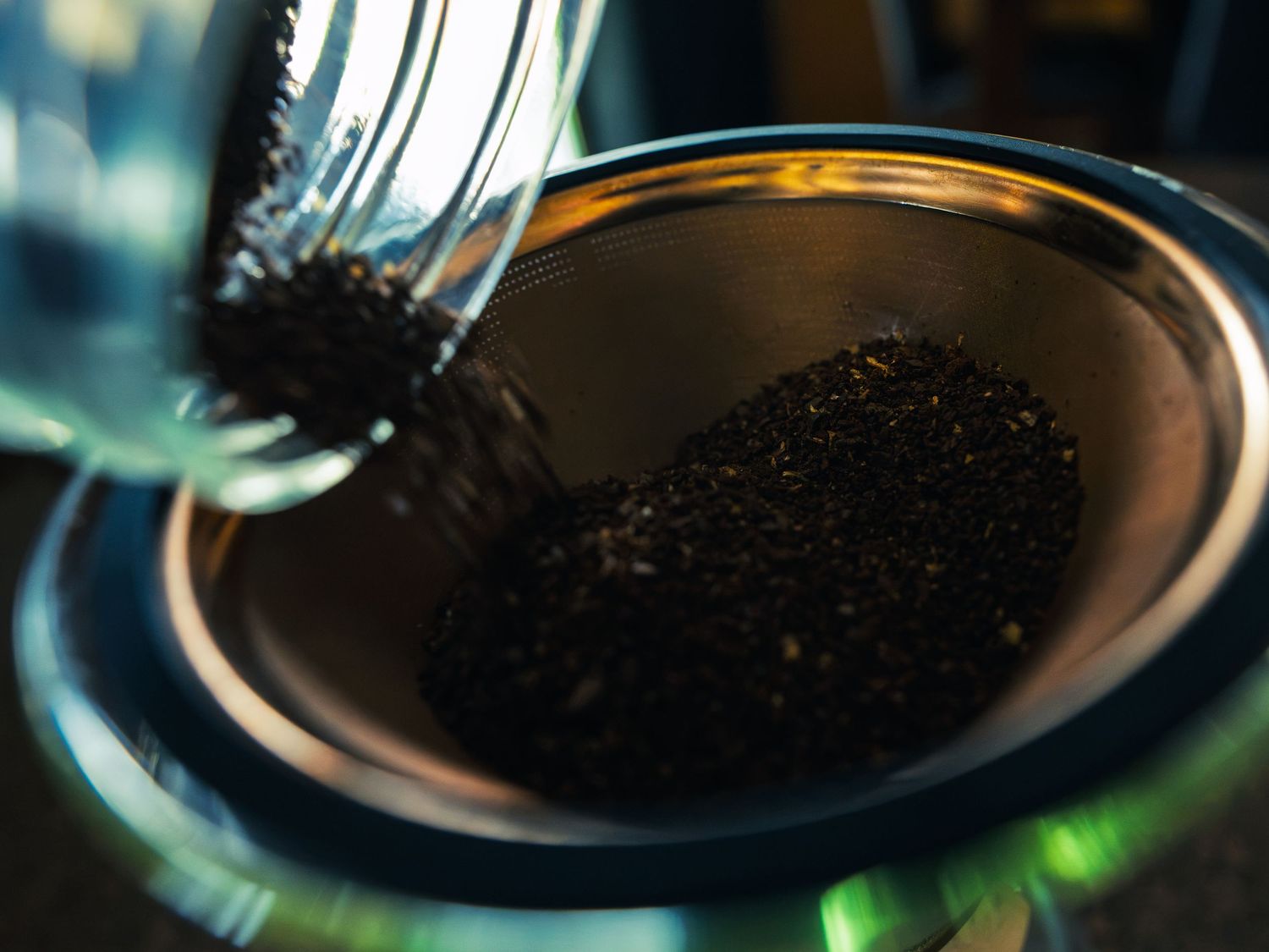 Freshly ground coffee pouring from a glass into a stainless steel pour-over filter, lit with warm and cool tones.
