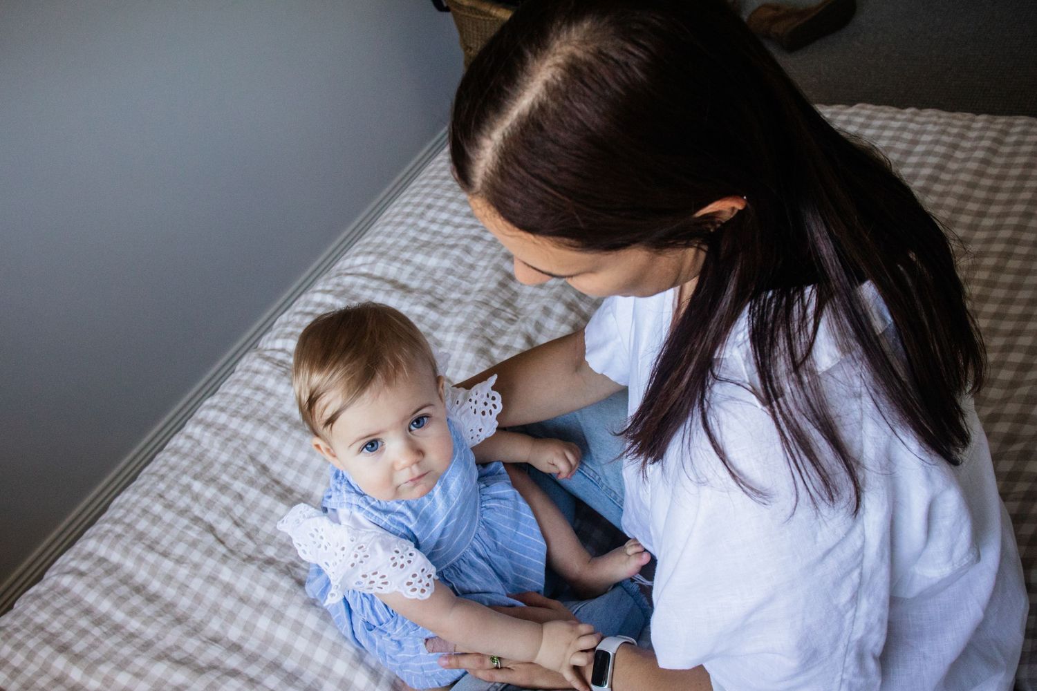 A Mother and baby in a blue dress sitting on a bed during an at-home mother and baby photoshoot in Baulkham Hills, Sydney.