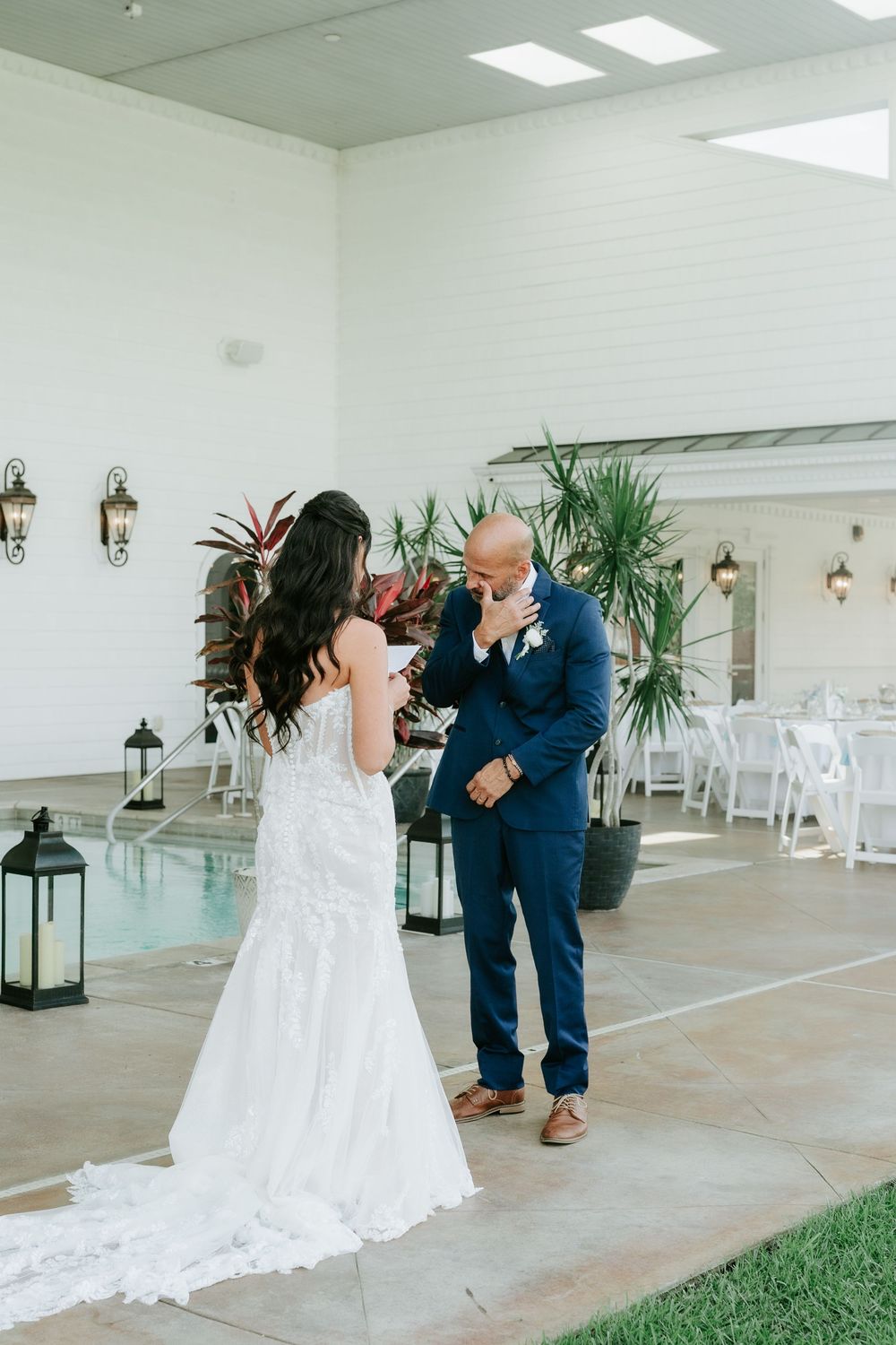 Bride in lace gown reads vows to emotional groom in navy suit during outdoor wedding ceremony near pool.
