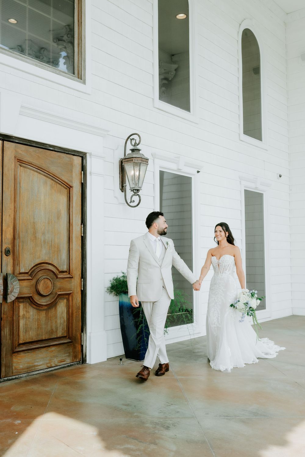 Bride and groom holding hands walking outside a white building with wooden doors on their wedding day.