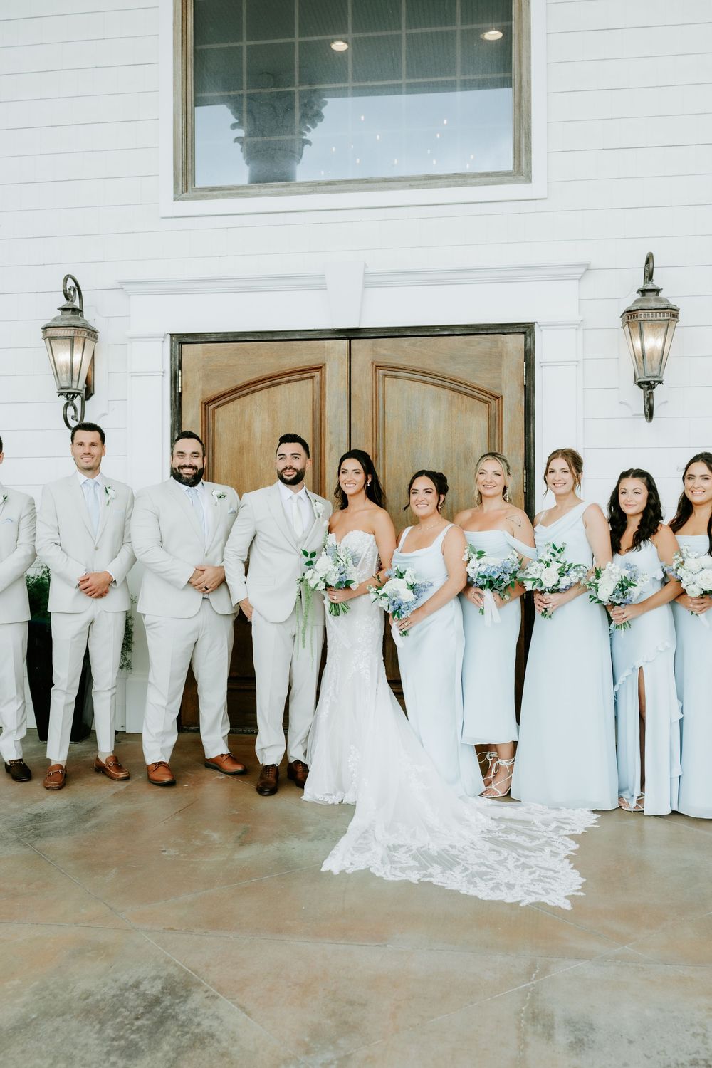 Wedding party poses outside white venue, groomsmen in tan suits and bridesmaids in light blue gowns holding white floral bouquets.
