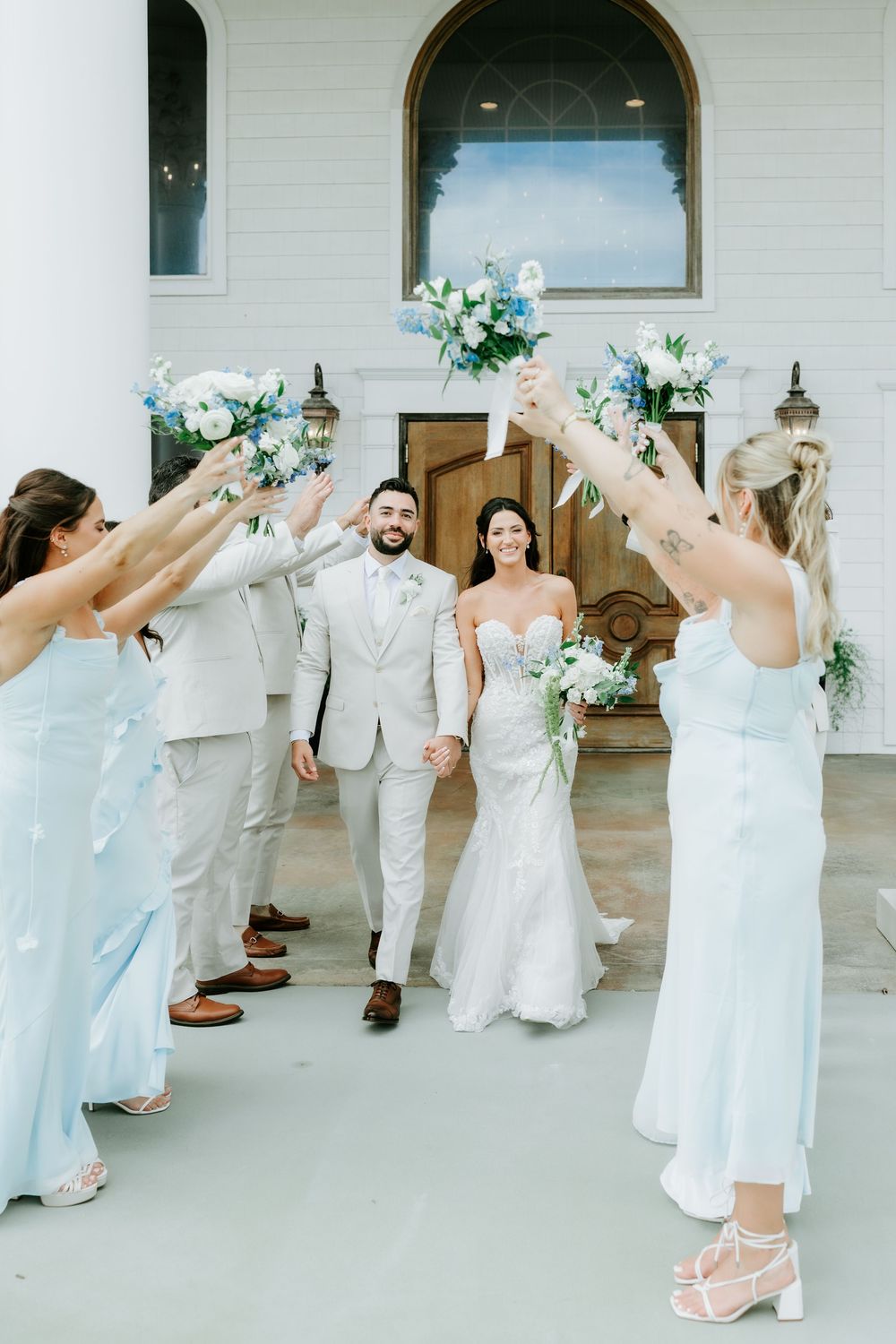 Bride and groom exit white chapel holding hands as wedding party raises blue floral bouquets in celebration.