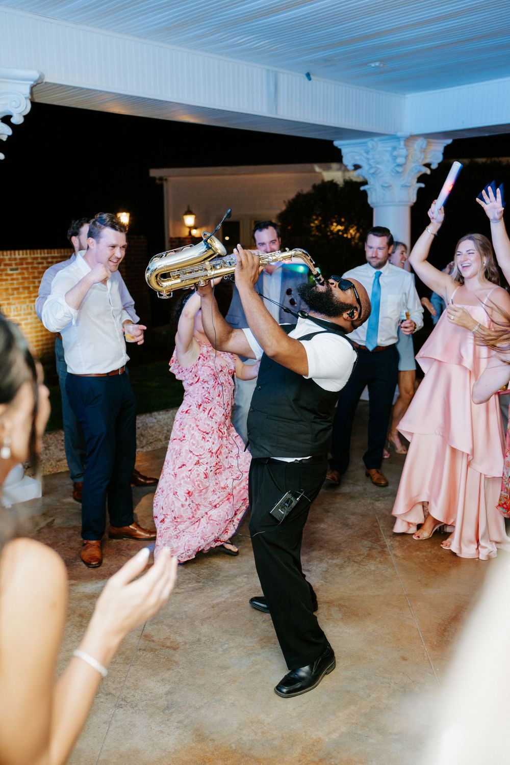 Saxophonist performing at lively outdoor wedding reception with guests dancing and celebrating at night.