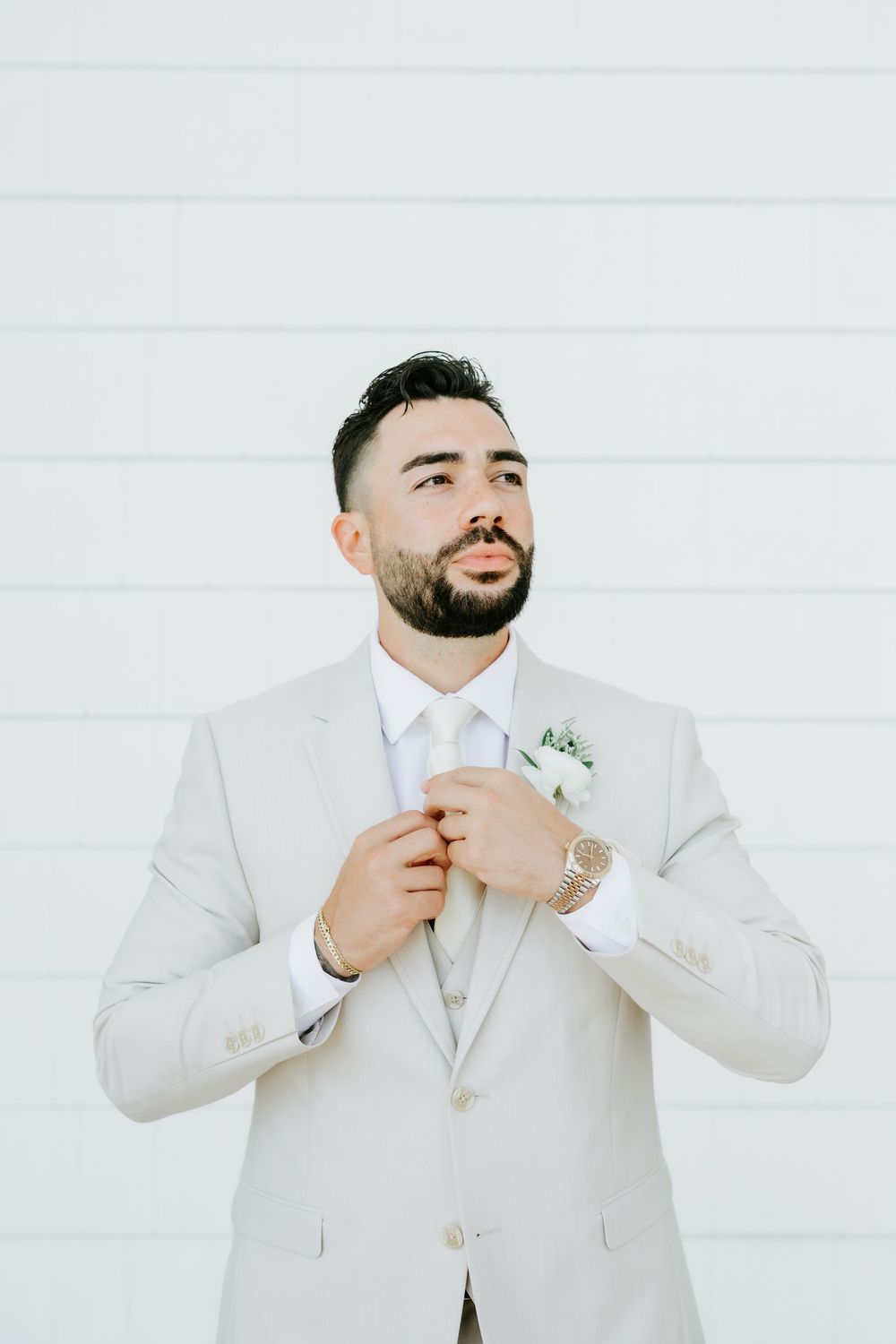 Groom in light gray suit adjusting tie with boutonniere, posing against white shiplap wall.