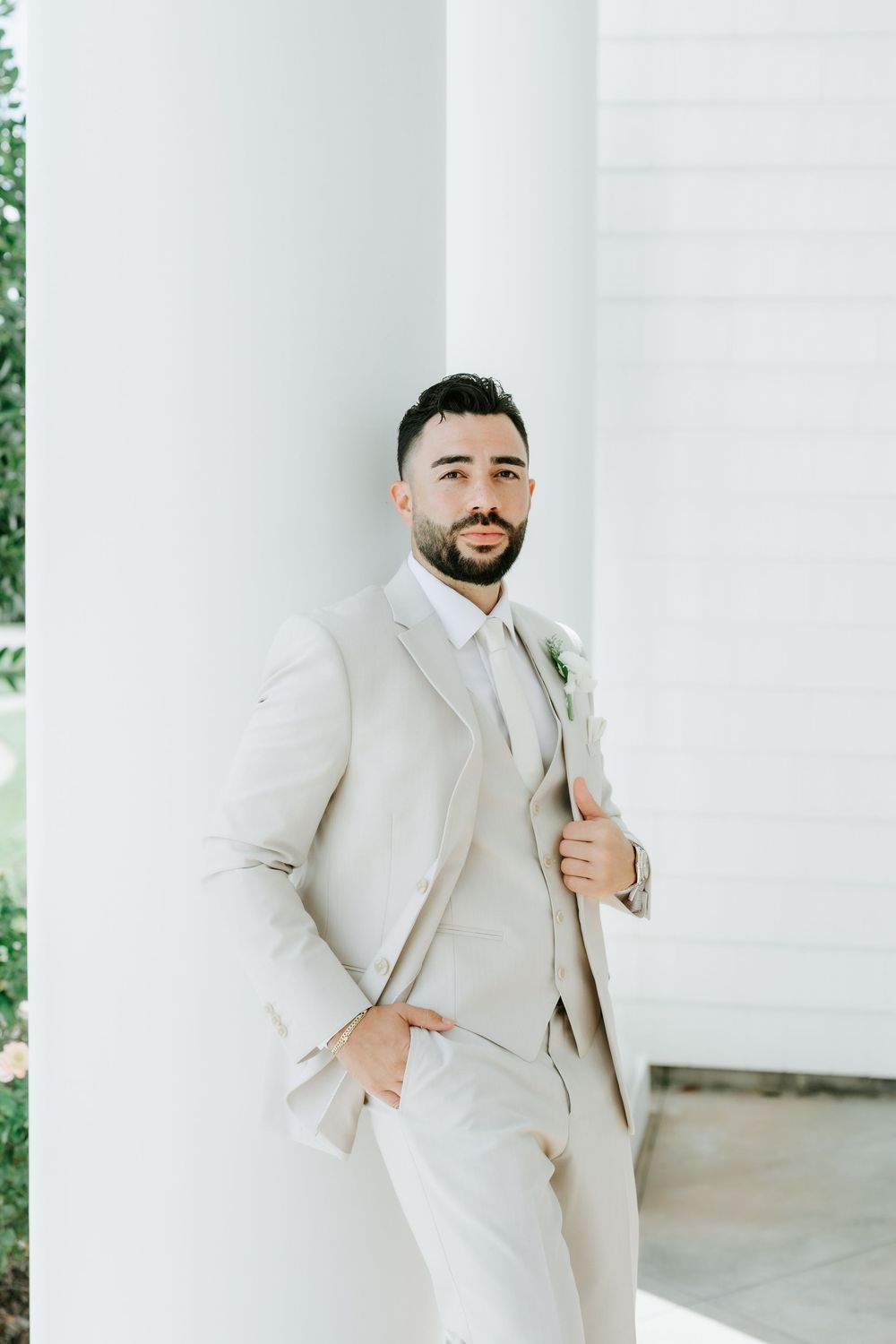 Groom in a beige three-piece suit leaning against a white column, holding lapel with one hand in pocket.