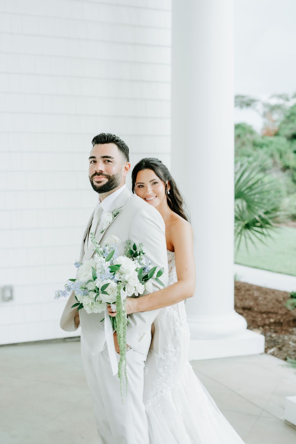 Bride and groom pose back-to-back at white colonial venue, bride holding white and blue bouquet in lace wedding gown.