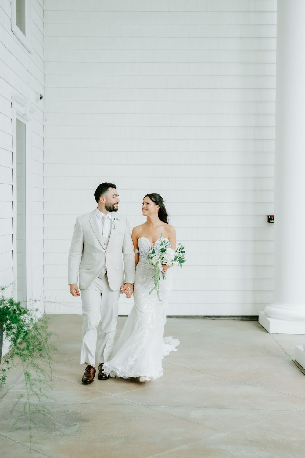Bride and groom walk hand-in-hand along white shiplap venue, bride in lace gown holding white floral bouquet.