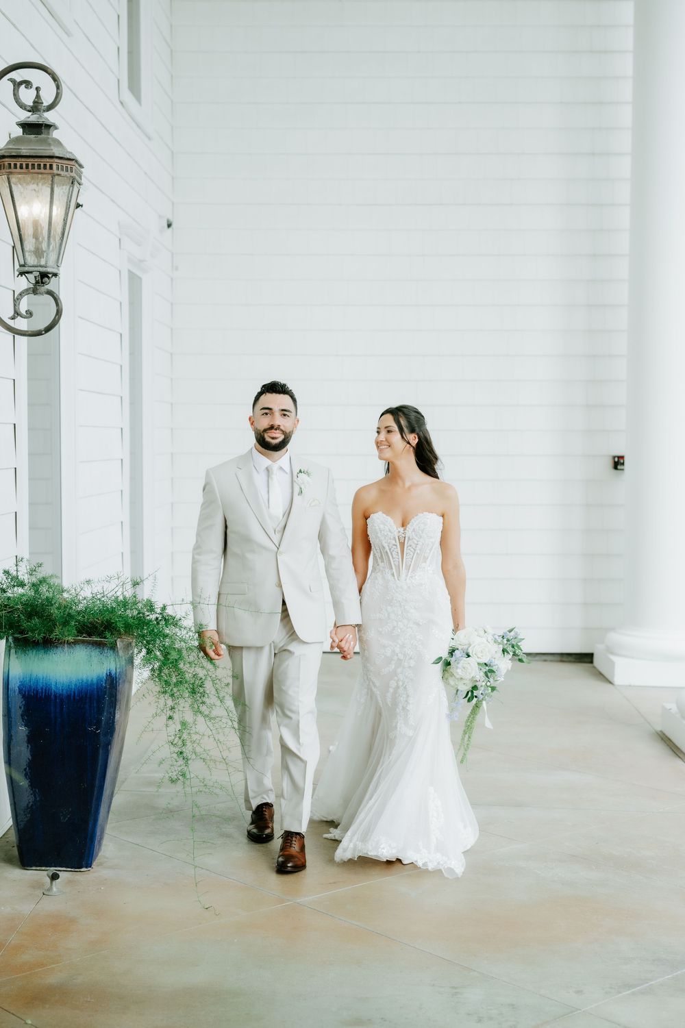 Smiling bride and groom hold hands walking past blue planter at elegant white Southern wedding venue.