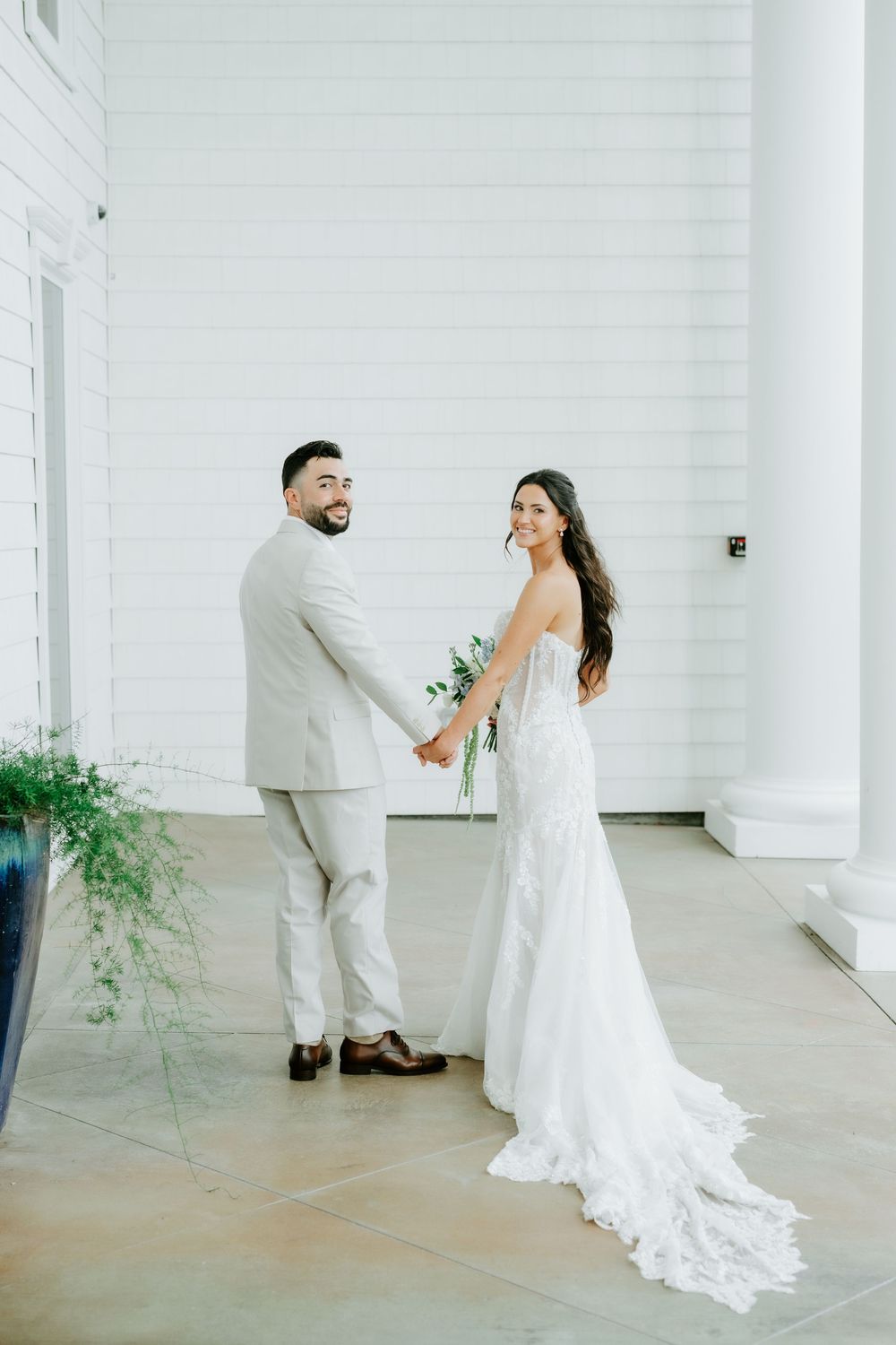 Bride and groom hold hands smiling in front of white building, bride in lace gown with train, groom in tan suit.
