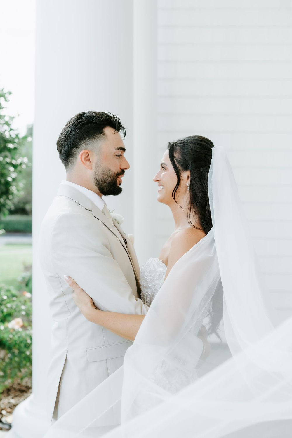 Bride and groom embrace and smile at each other outdoors, with flowing veil and white columns backdrop.