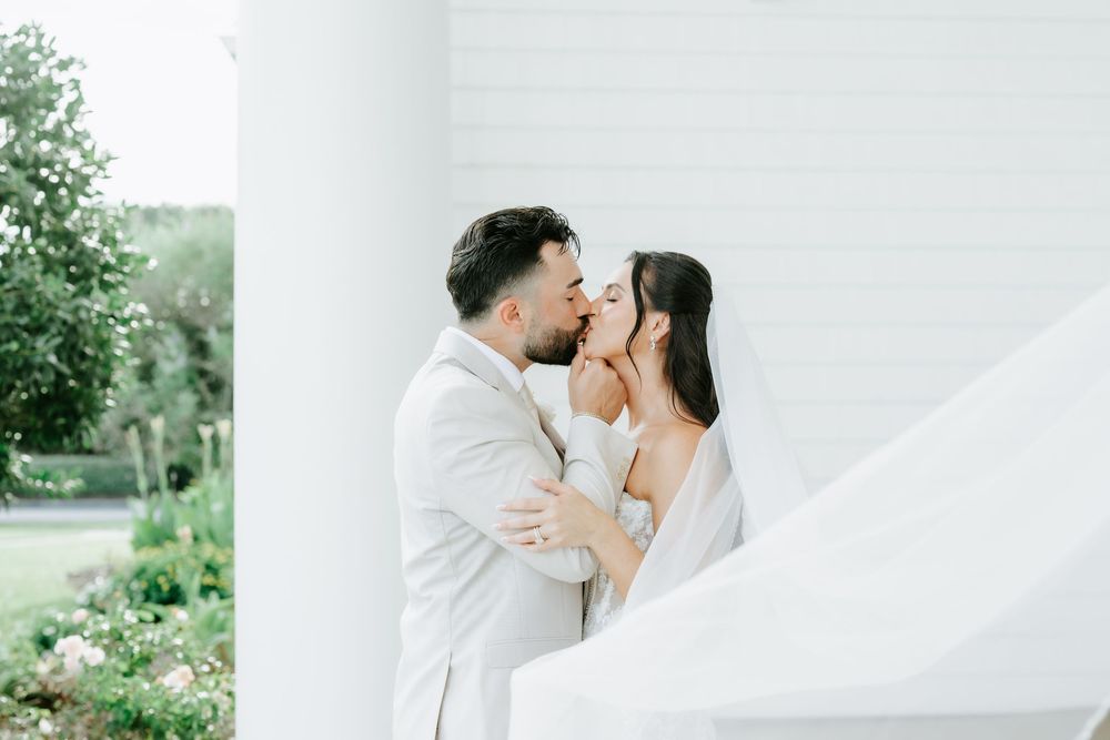 A bride and groom share a romantic kiss outdoors near white columns, with the bride's veil flowing in the breeze.
