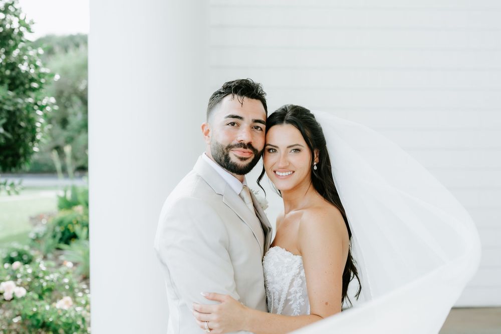 Happy couple posing outdoors on their wedding day, groom in white suit, bride in strapless gown with veil flowing.