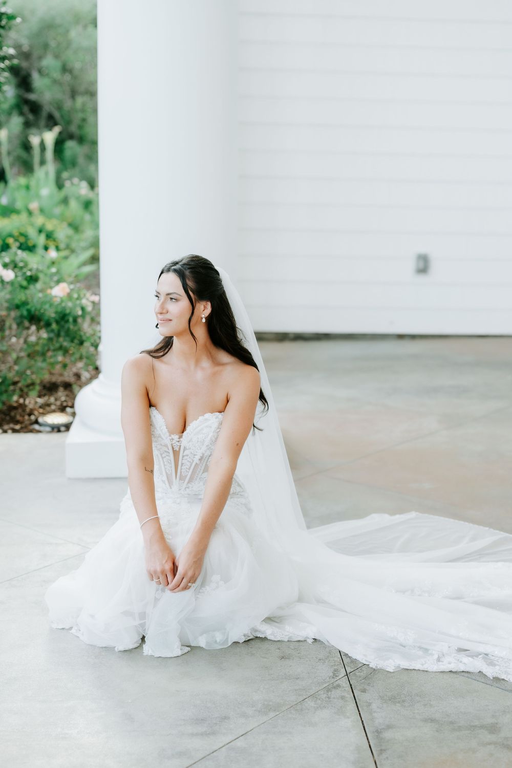 Elegant bride in strapless lace wedding gown sits gracefully on floor beside white column with veil trailing.