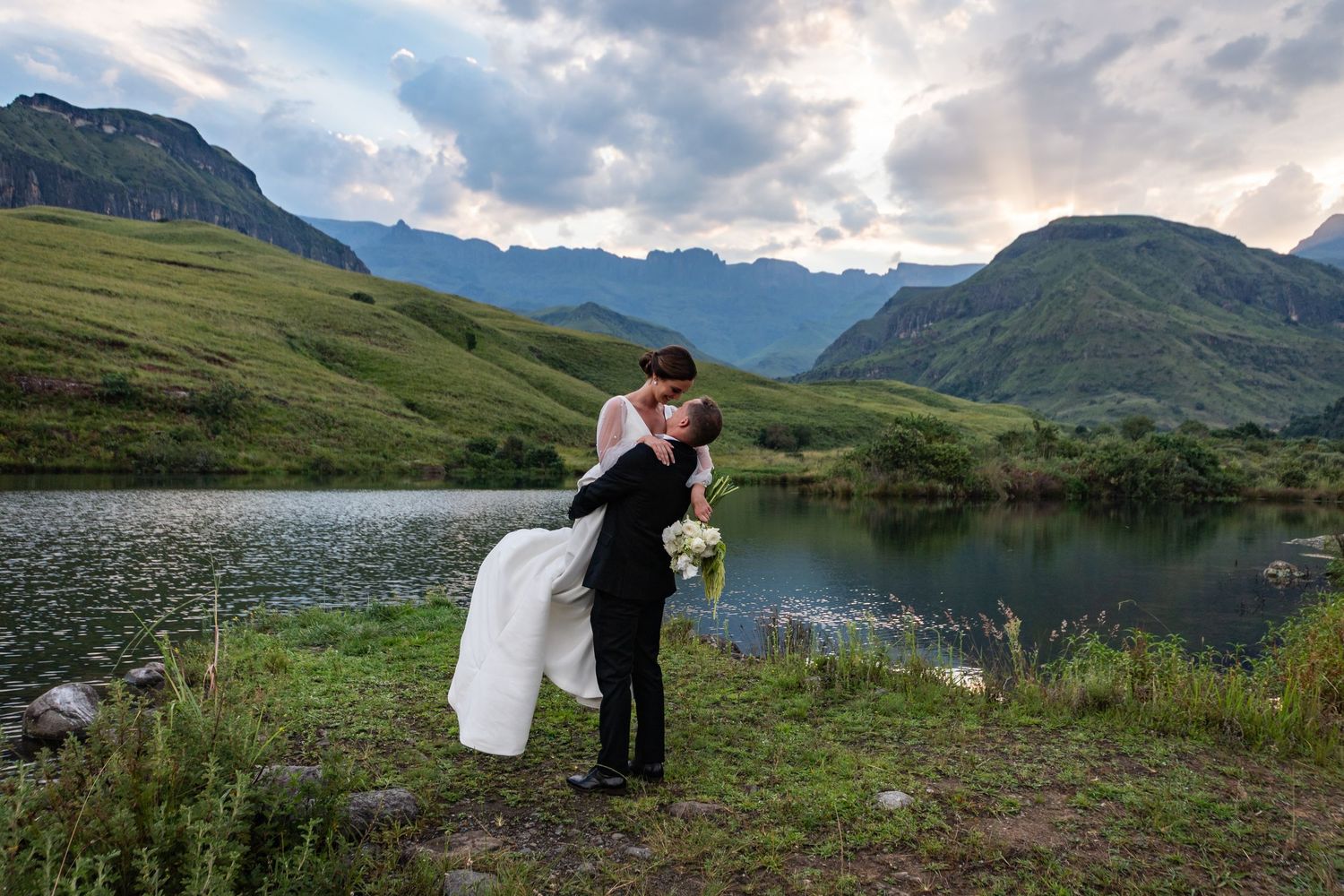 Bride and groom share a romantic kiss by a mountain lake with lush green hills and dramatic cloudy skies.