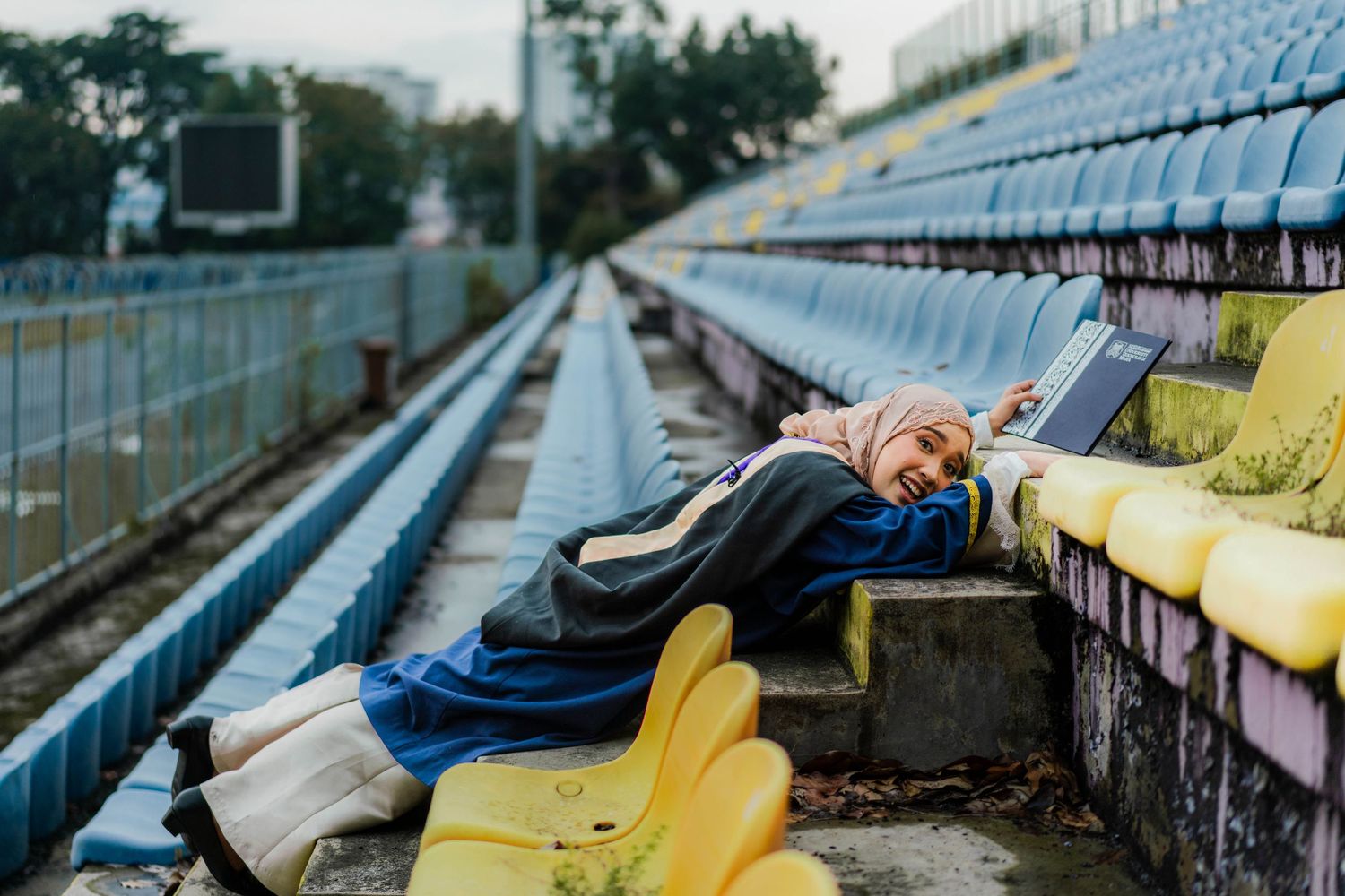 Woman in hijab lying on stadium bleachers using a laptop computer outdoors.