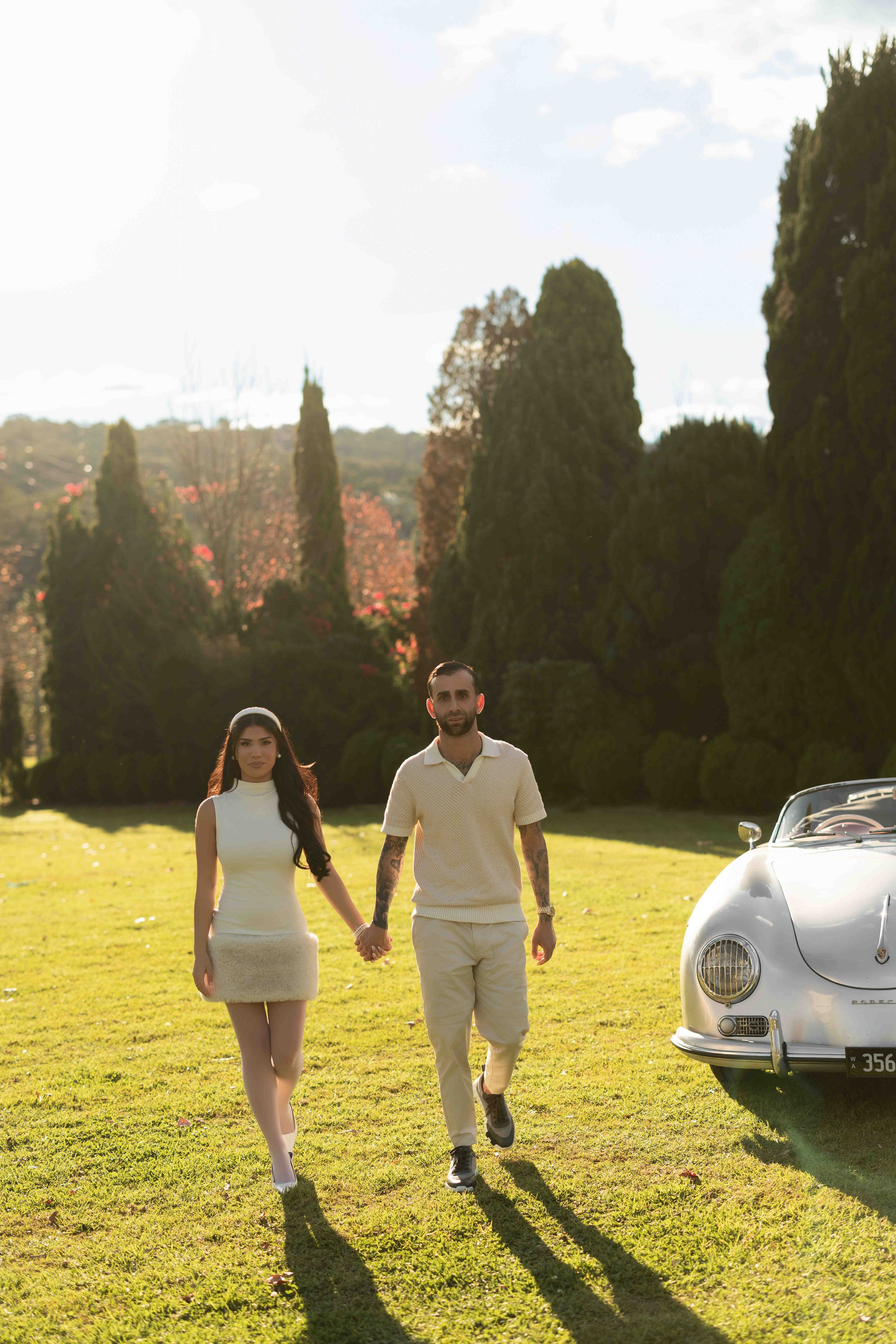 Couple holding hands walking on green grass beside a vintage Porsche 356 convertible in a sunny park.