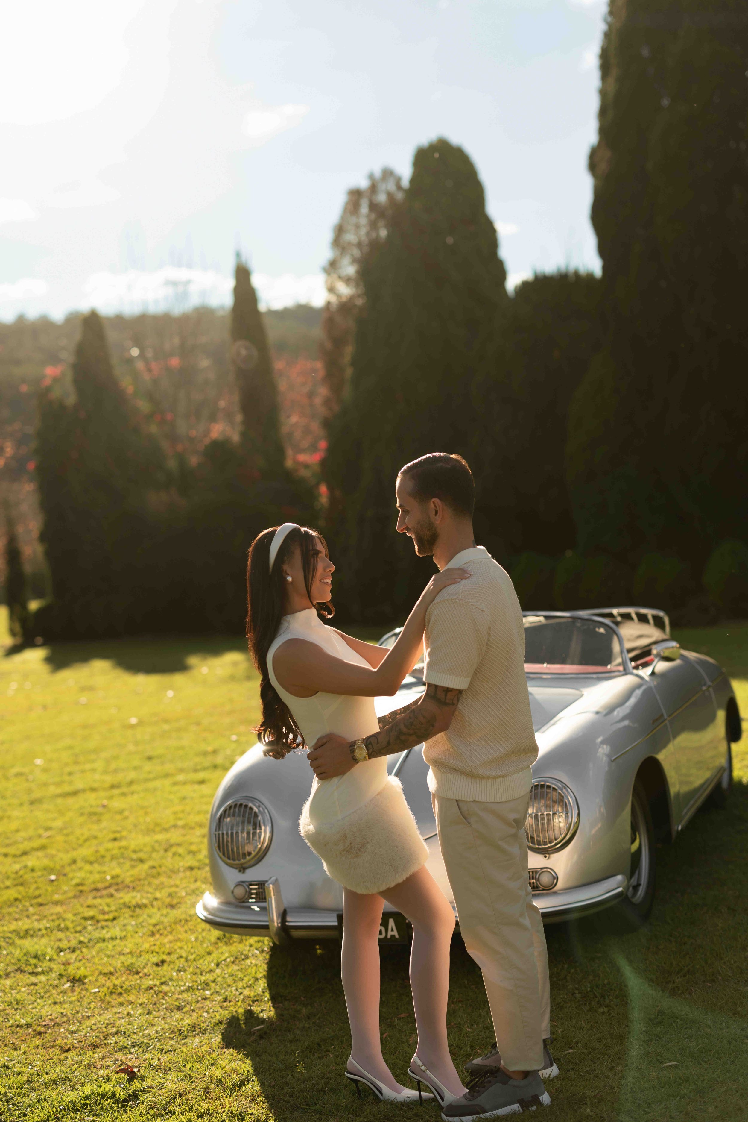 Couple embracing beside a vintage white convertible car on a sunny green lawn with tall cypress trees in the background.