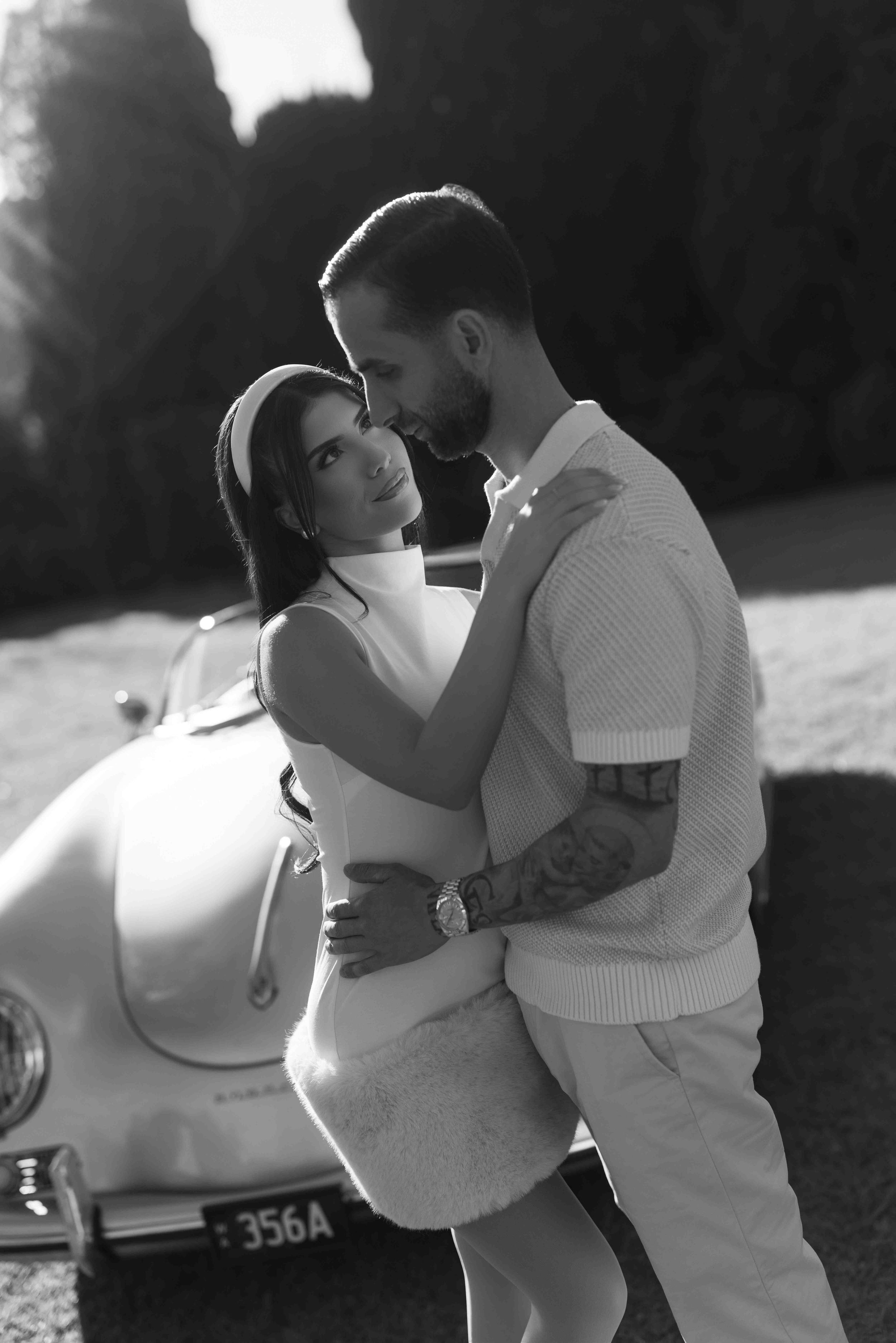 Black and white photo of a couple embracing romantically in front of a vintage classic car outdoors.