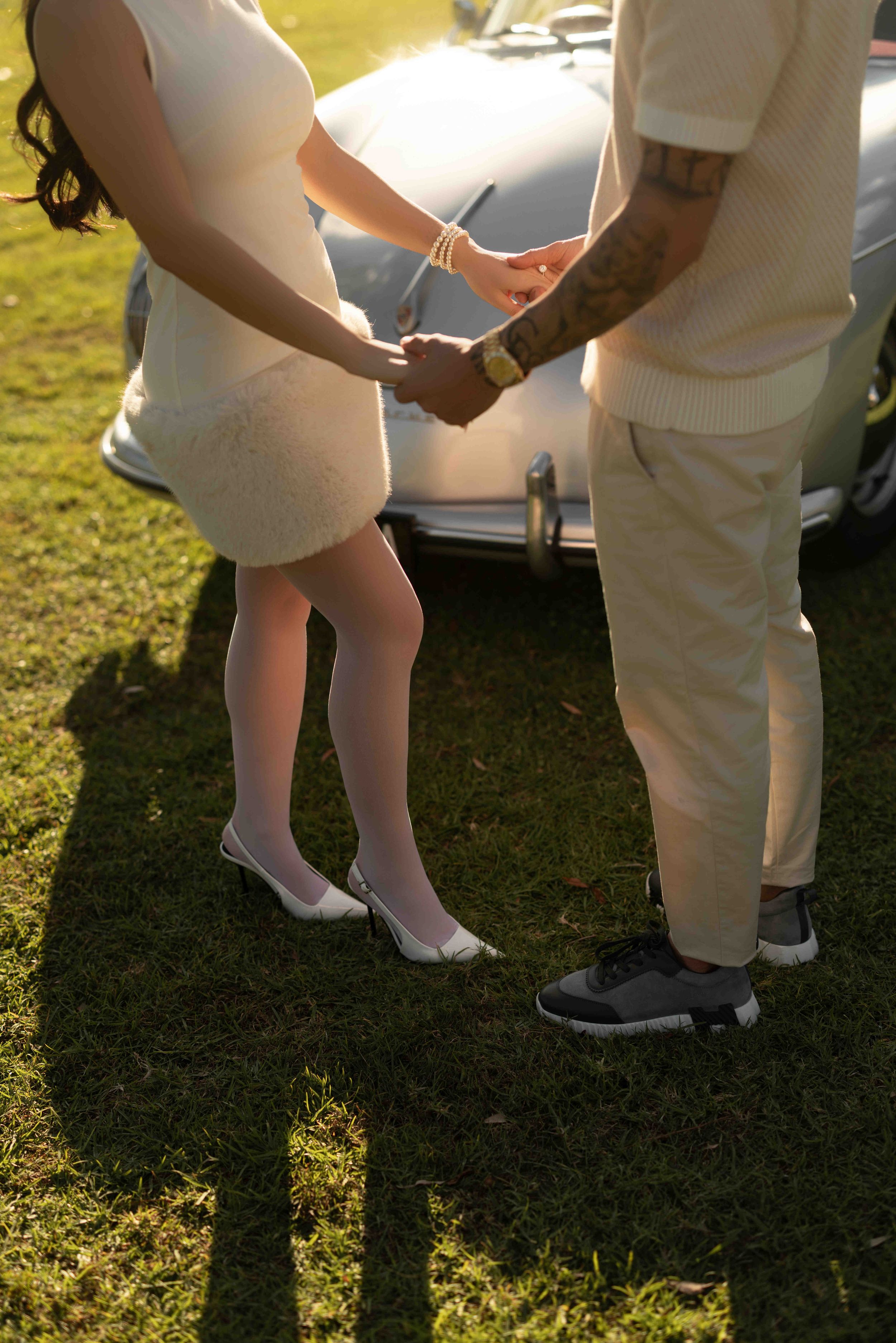 Couple holding hands in matching cream outfits standing on grass near a vintage car in golden sunlight.
