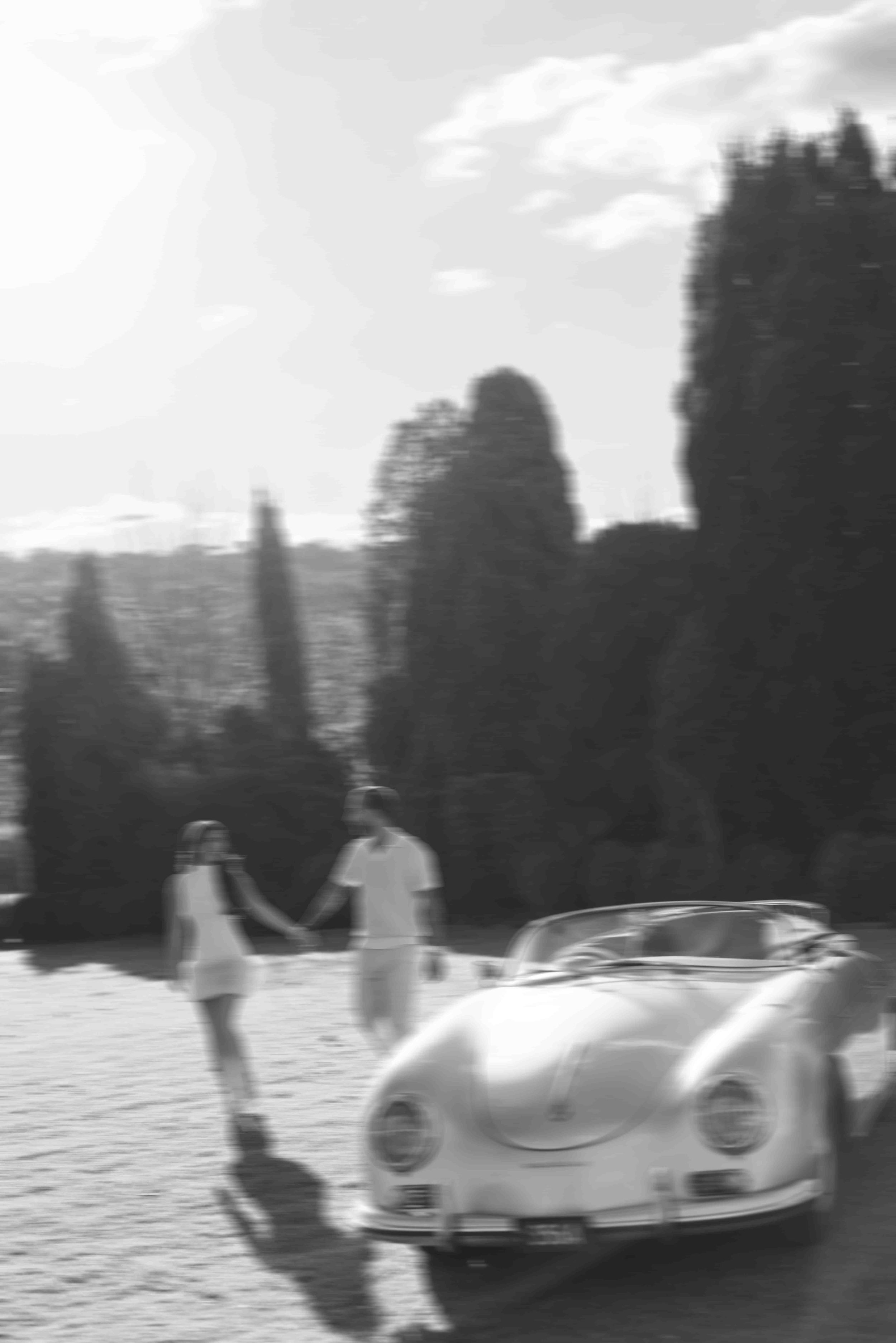 Black and white photo of a couple holding hands beside a vintage Porsche convertible on a sunny tree-lined road.