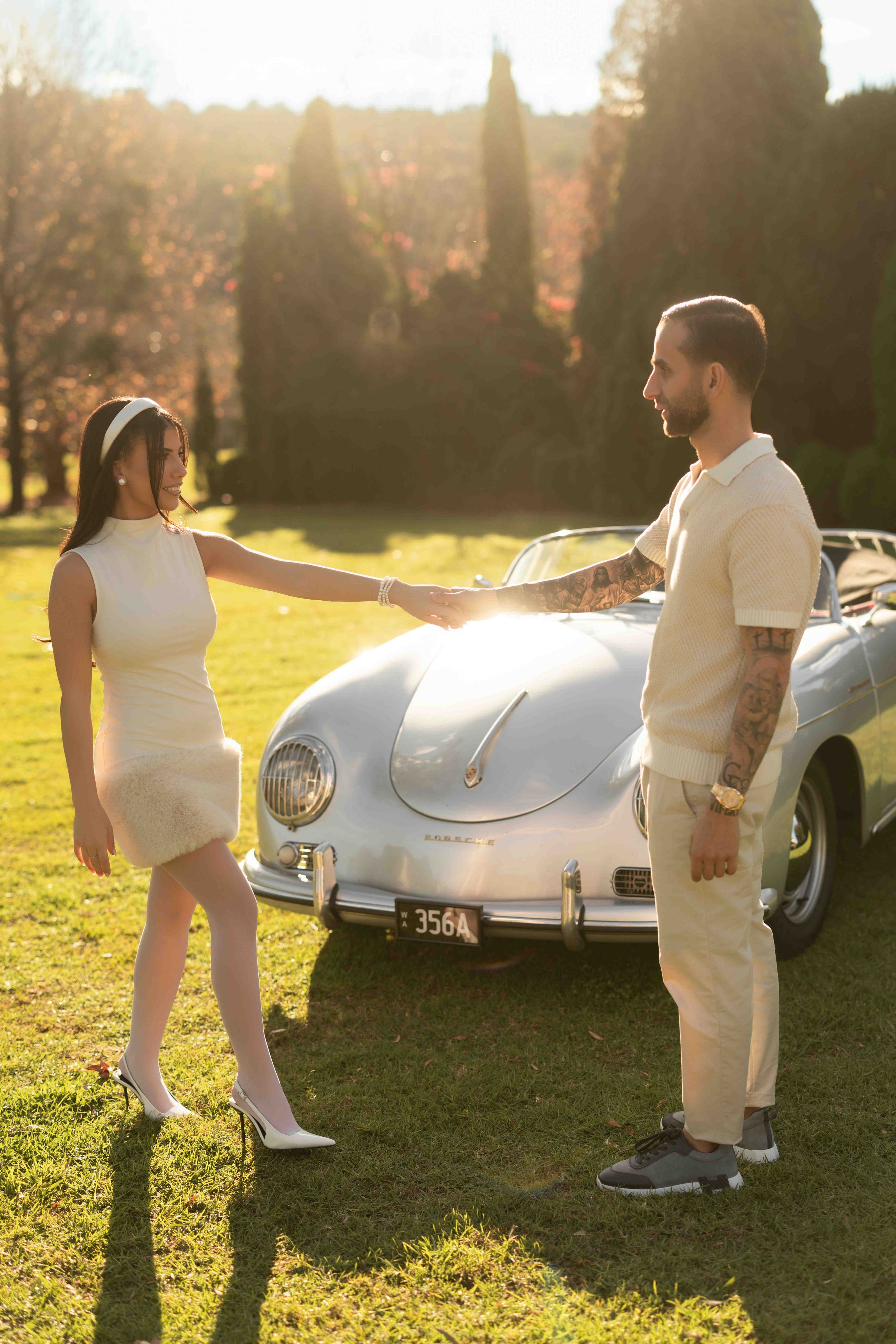 Couple holding hands by a vintage Porsche 356 convertible in golden sunset light, woman in white dress, man with tattoos.