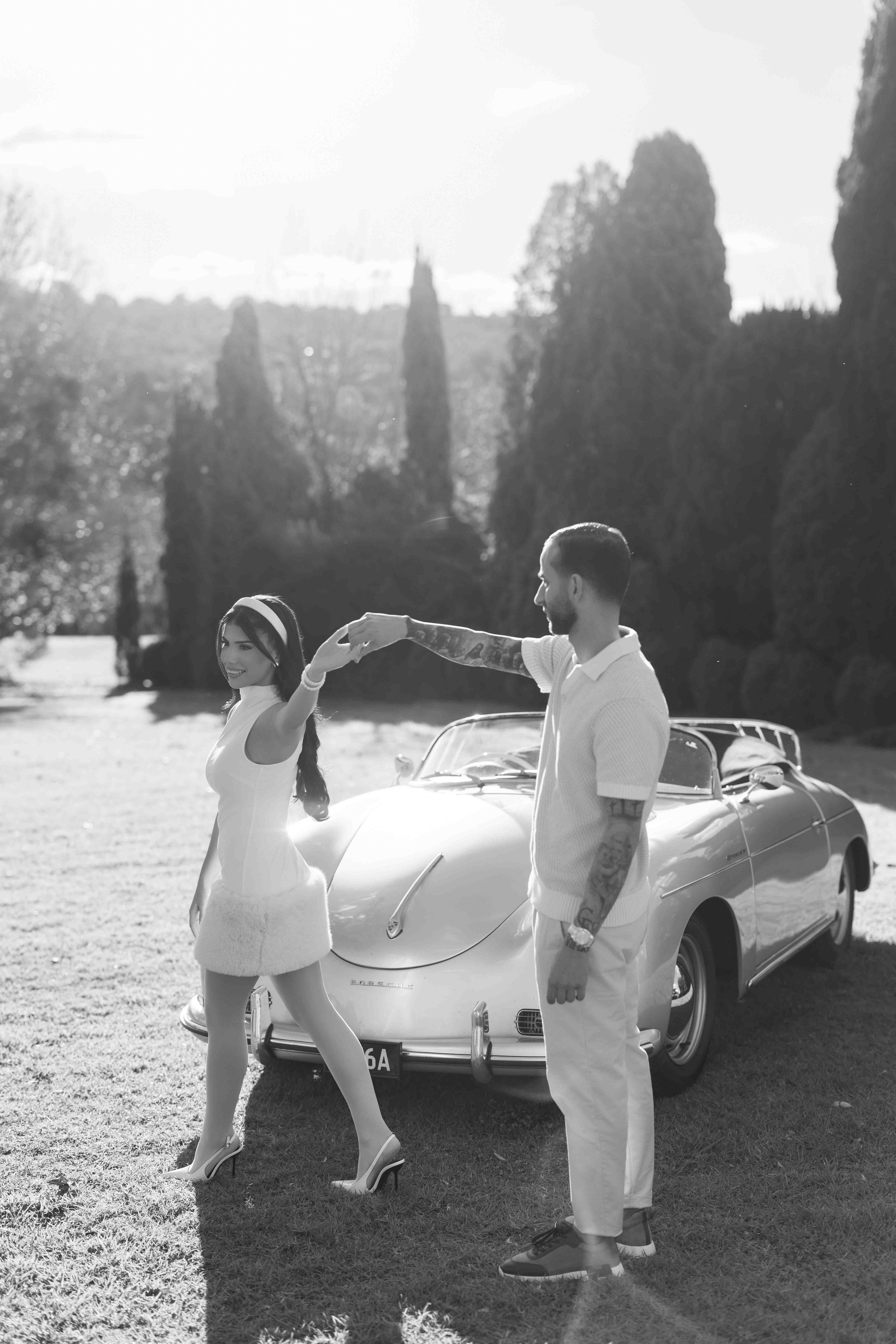 Black and white photo of a couple dancing beside a vintage convertible car surrounded by cypress trees.