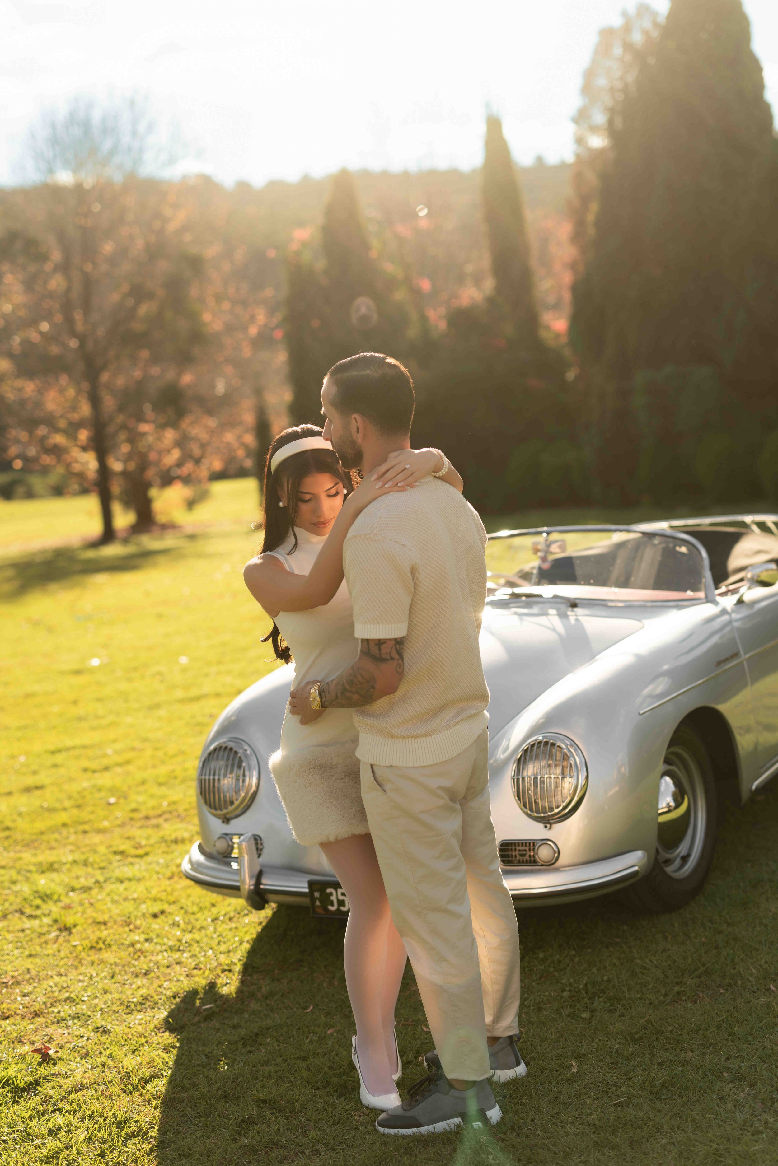 Couple embracing romantically beside a vintage silver Porsche convertible in a sunlit autumn park setting.