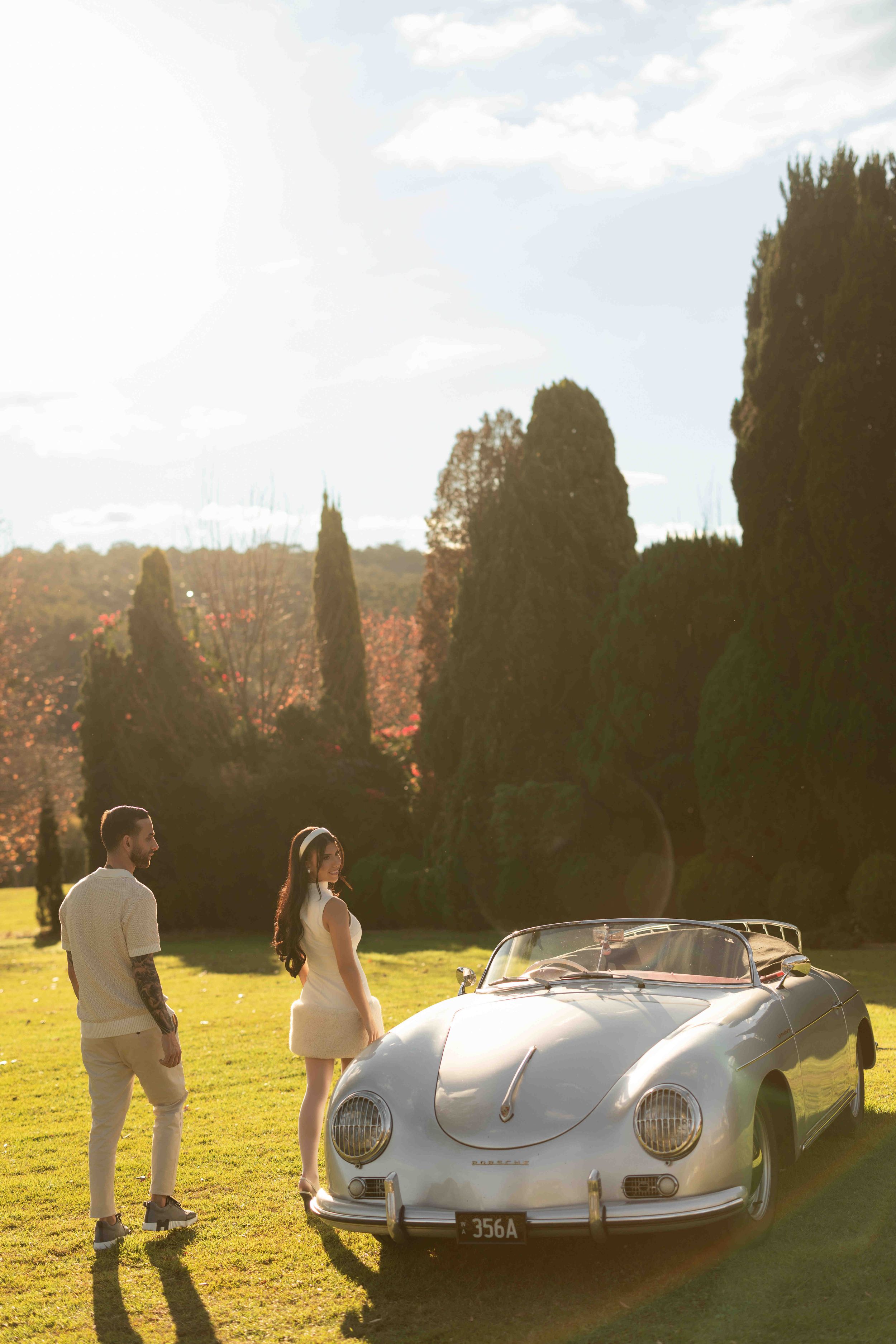 Couple standing beside a vintage white Porsche 356A convertible on a sunlit lawn surrounded by tall cypress trees.