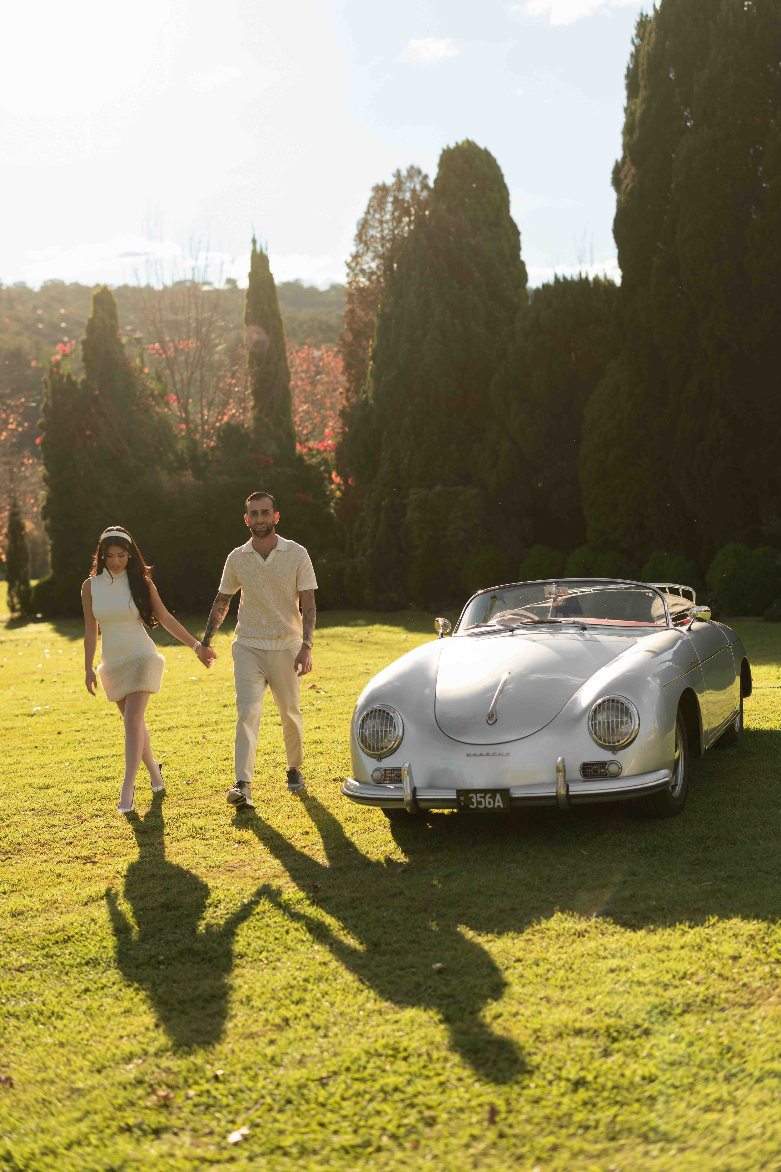 Couple in white outfits holding hands on green lawn beside a classic white Porsche convertible with tall cypress trees.