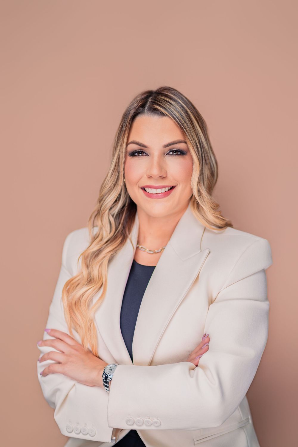 Professional woman in white blazer smiling with arms crossed against a neutral beige background.