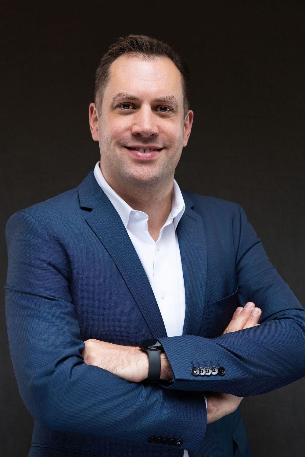 Professional businessman in blue blazer and white shirt smiling with arms crossed against dark background.