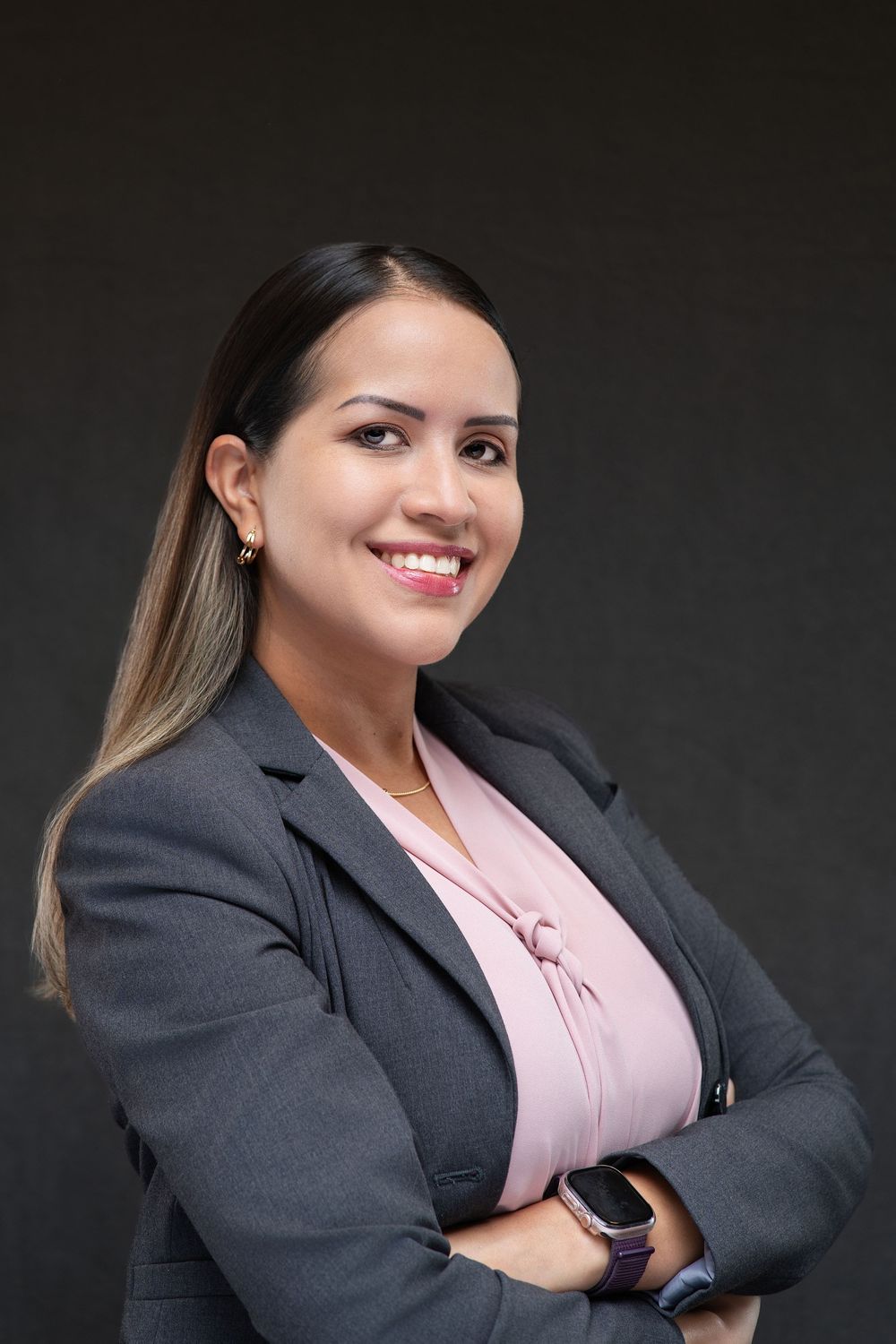 Professional businesswoman in gray blazer and pink blouse smiling with arms crossed against dark background.
