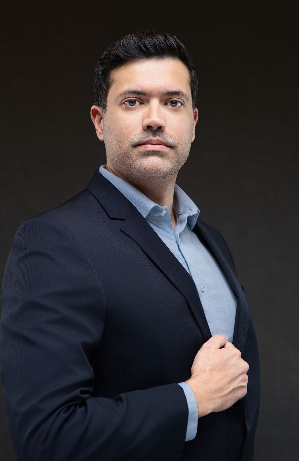 Professional man in navy blazer and light blue shirt posing confidently against dark background.