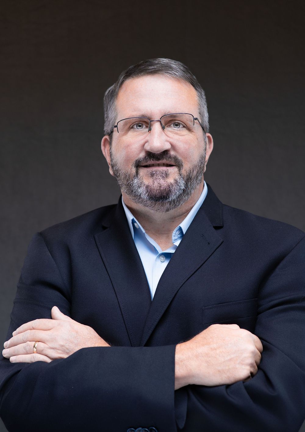 Professional man in black blazer with crossed arms, wearing glasses and beard, posing confidently against dark background.