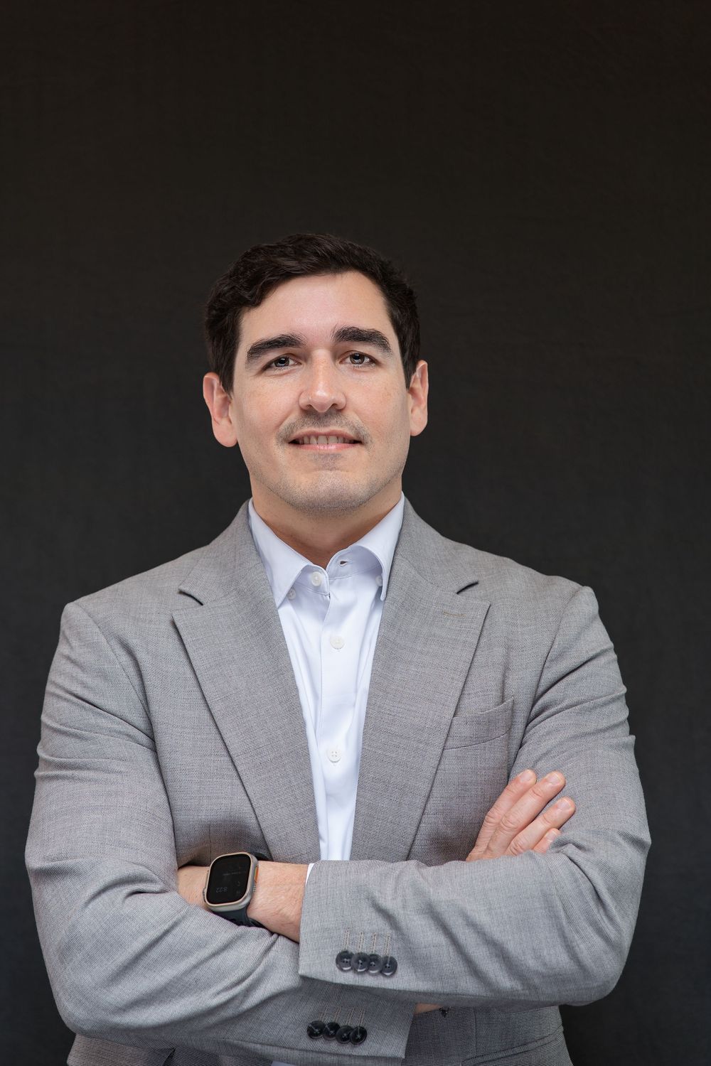 Professional businessman in gray suit with arms crossed smiling confidently against dark background.